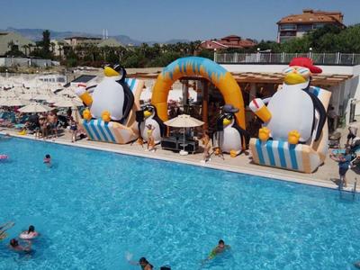 Una grande piscina di un hotel con decorazioni gonfiabili a forma di pinguino e ospiti che nuotano sotto un cielo blu.