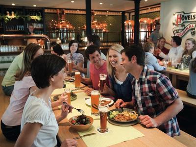 Grupa młodych ludzi siedzi przy drewnianym stole w restauracji, ciesząc się posiłkami i napojami.