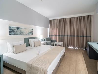 Jasny pokój hotelowy z dużym łóżkiem podwójnym, biurkiem i częścią wypoczynkową obok dużych zasłon.