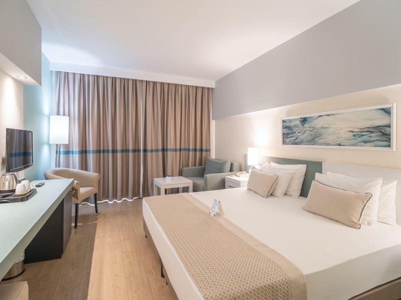 Nowoczesny pokój hotelowy z dużym łóżkiem, telewizorem i biurkiem w neutralnych kolorach.