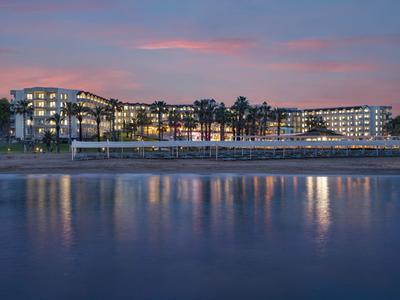 Grand bâtiment d'hôtel avec fenêtres éclairées au bord d'une eau calme au coucher du soleil.