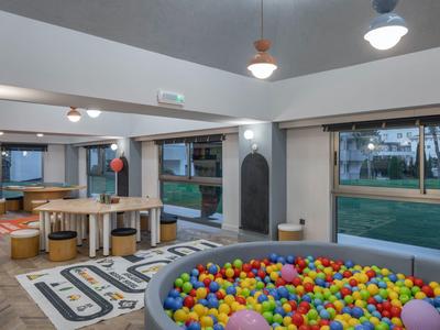 Salle de jeux pour enfants avec piscine à balles, tables, canapés, grandes fenêtres et vue extérieure.