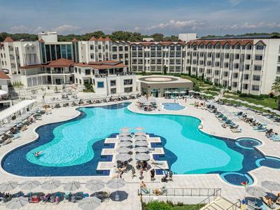 Large hotel pool with sun loungers and umbrellas in a complex with multi-story buildings.