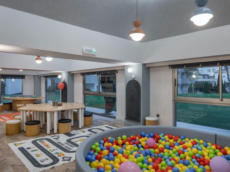 Salle de jeux pour enfants avec piscine à balles, tables, canapés, grandes fenêtres et vue extérieure.