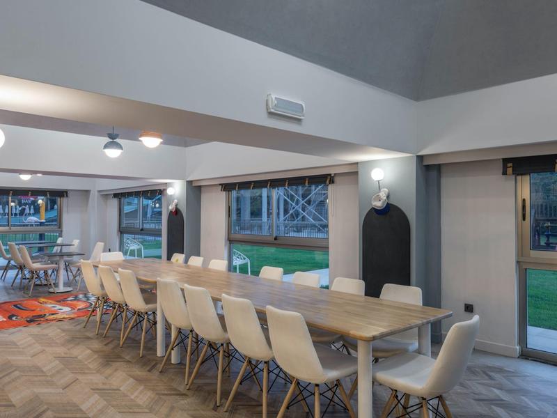 Salle à manger moderne avec grande table en bois et chaises blanches, près de grandes fenêtres donnant sur l'extérieur.