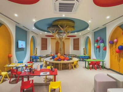 Area giochi colorata per bambini con tavoli, sedie, decorazioni alle pareti e un albero al centro.