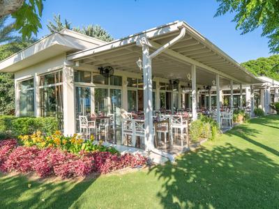 Nowoczesna biała restauracja ze szklanymi panelami otoczona ogrodem i drzewami w słoneczny dzień.