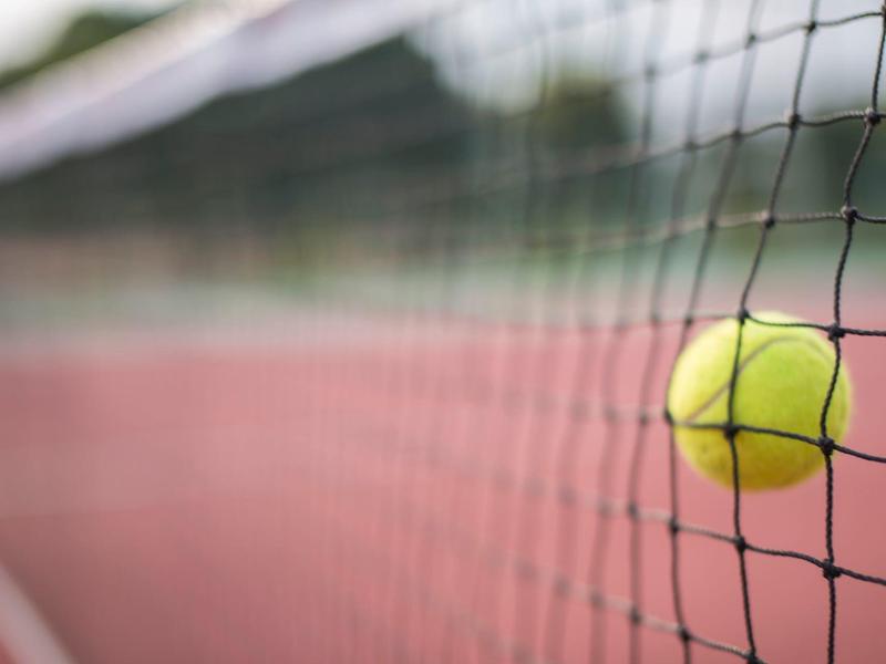 Piłka tenisowa zawieszona w siatce na czerwonym korcie tenisowym pod zachmurzonym niebem.