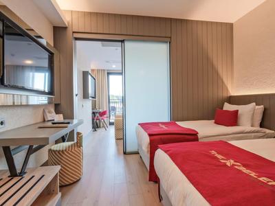 Habitación de hotel moderna con dos camas, ropa de cama roja y acceso a una habitación contigua.