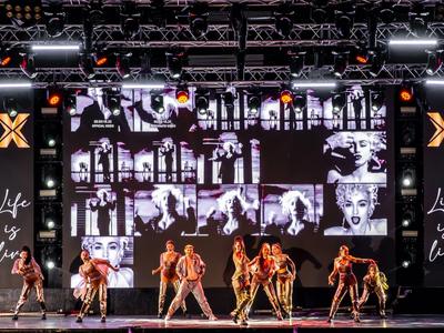 Gruppo di danza che si esibisce su un palco con grande schermo video e illuminazione.