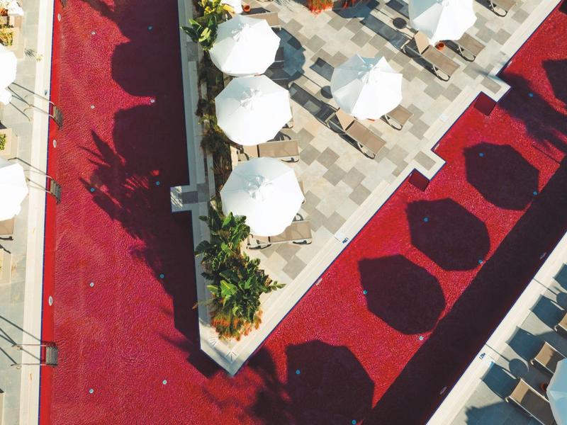 Vista aérea de una piscina de hotel con baldosas rojas y sombrillas blancas alrededor.