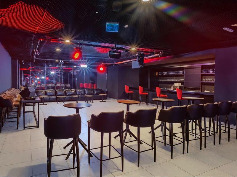 Área de bar moderno en hotel con taburetes altos y luces rojas.