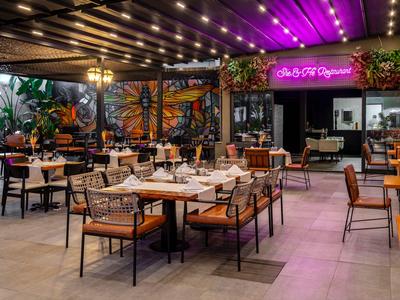 Modern restaurant with wooden furniture, plants, and purple lighting in an indoor courtyard.