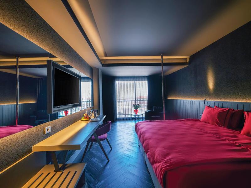 Chambre d'hôtel moderne avec grand lit, literie rouge, bureau et balcon avec vue.