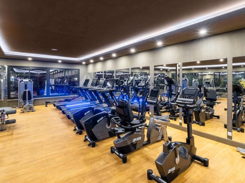 Une salle de sport moderne avec sol en bois, miroirs et divers équipements sous un éclairage au plafond.