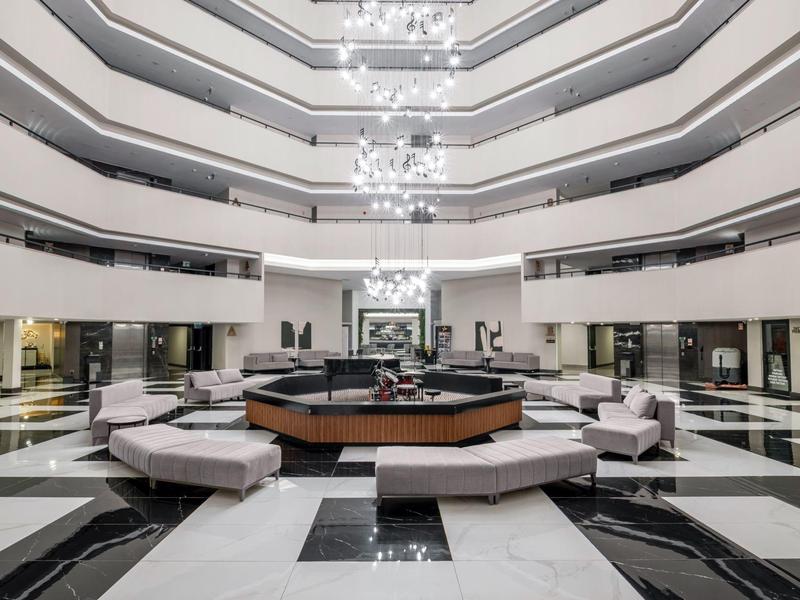 Grande hall d'hôtel avec mobilier moderne, espaces assises et fontaine centrale sous des lustres.