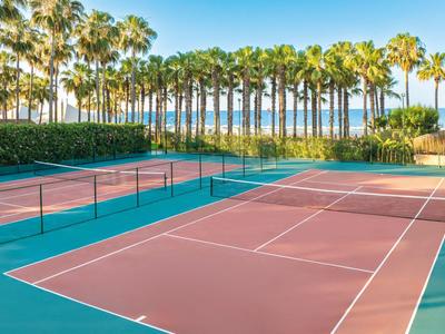 Un campo da tennis con campi rossi e verdi, circondato da palme con vista sul mare.