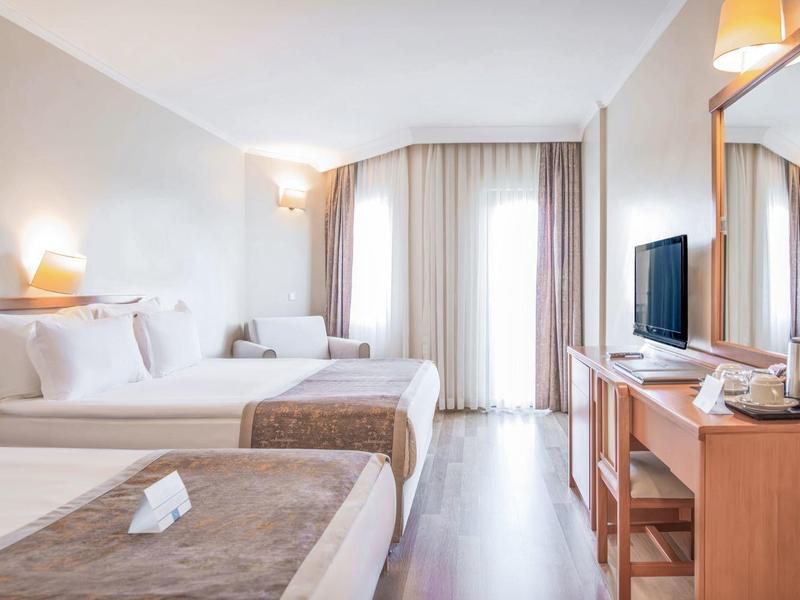 Camera d'hotel moderna con due letti singoli, scrivania, TV e mobili in legno chiaro.