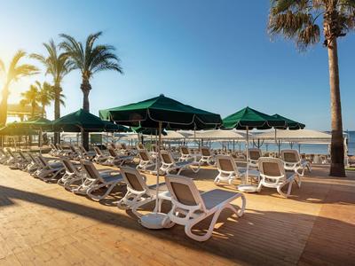 Sunlit poolside with empty white loungers and green umbrellas by the sea under palm trees.