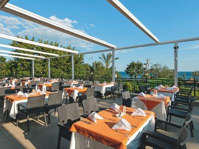 Outdoor restaurant seating with orange tablecloths and folded napkins under a pergola near greenery.