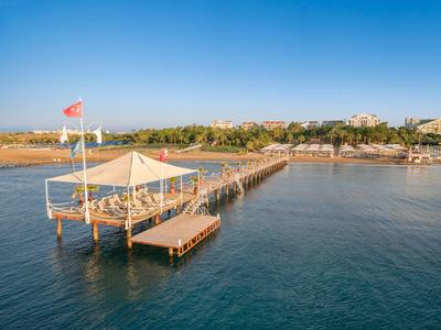 Wooden pier with shaded seating extends over calm blue sea under clear sky near coastline.