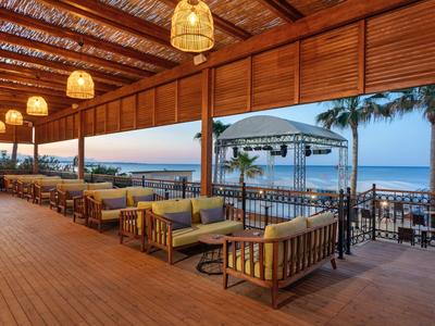 Outdoor beachside lounge with wooden furniture, hanging lights, and a view of the ocean at sunset.