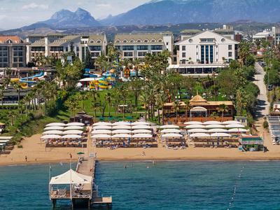 Beachfront hotel with umbrellas, pier, palm trees, and mountain backdrop under clear sky.
