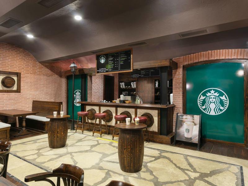 Cozy Starbucks cafe interior with wooden furniture, stone floor, and Starbucks logo on green walls.