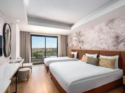 Chambre d'hôtel moderne avec deux lits, bureau, télévision et balcon offrant une vue sur un paysage verdoyant.