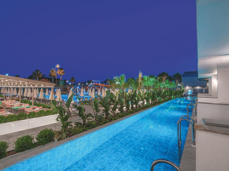 Une zone de piscine éclairée au crépuscule à côté d'une terrasse d'hôtel avec des plantes.