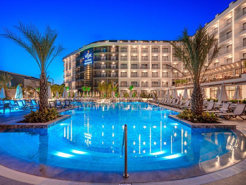 Grand hôtel avec piscine extérieure illuminée et palmiers au crépuscule.