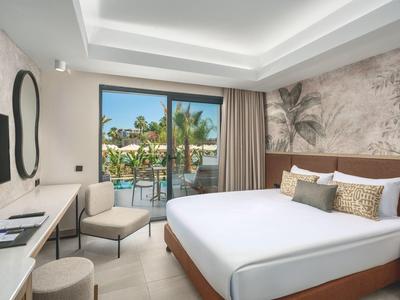 Hotelkamer met groot bed, balkon met uitzicht op zwembad en palmbomen, modern interieur en zachte verlichting.