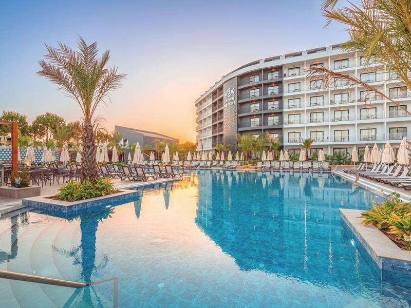 Modern hotel met groot buitenzwembad, ligstoelen en palmbomen bij zonsondergang.