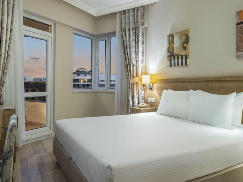 Ruime hotelkamer met tweepersoonsbed, bureau, tv en uitzicht op de stad door grote ramen.