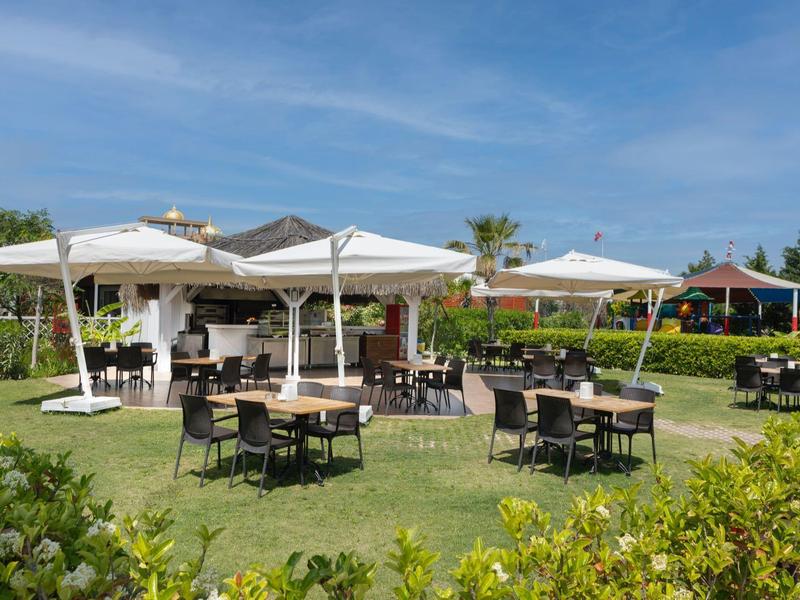 Buitenrestaurant met tafels, stoelen en parasols op groen gazon onder blauwe lucht.