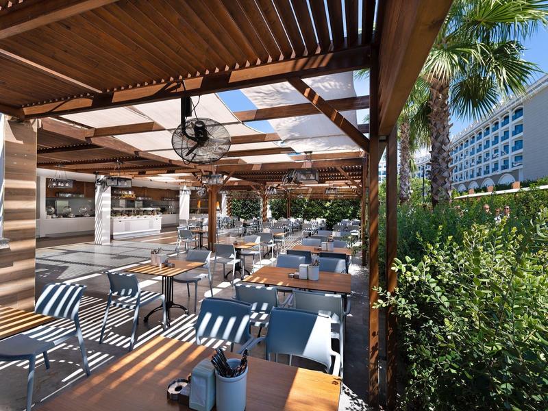 Overdekt terras met houten balken, ventilatoren aan het plafond, tafels, stoelen en omringend groen.