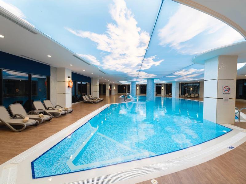 Moderní vnitřní bazén s lehátky a osvětleným stropem napodobujícím modrou oblohu.