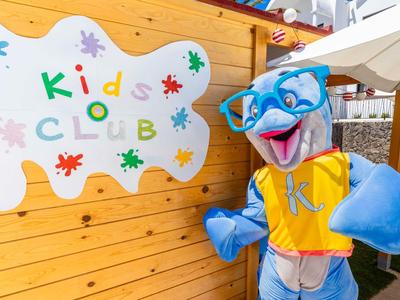 Maskottchen in knallgelb-blauem Kostüm vor bunter 'Kids Club'-Wand im Außenbereich.