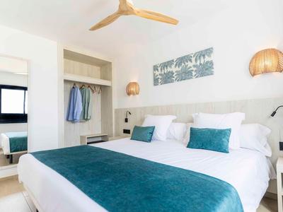 Hell eingerichtetes Hotelzimmer mit großem Bett, blauen Kissen und Decken, modernen Lampen und Deckenventilator.