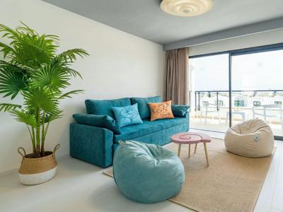 Modernes Wohnzimmer mit blauem Sofa, Sitzsäcken, grünem Pflanzen und großen Fenstern mit Balkonblick.