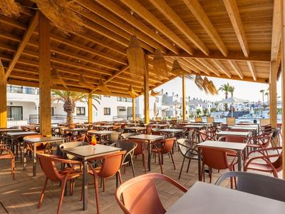 Überdachte Terrasse mit Holzdach, zahlreichen Tischen und Stühlen, umgeben von Gebäuden und Sonne.
