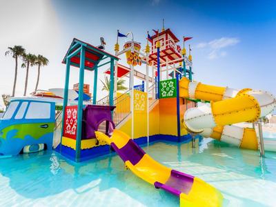 Buntes Wasserspiel mit Rutschen, Türmen und Palmen in klarem blauem Wasser unter blauem Himmel.