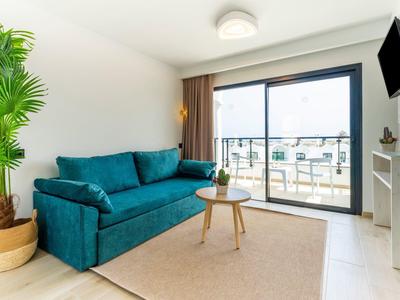 Helles Wohnzimmer mit blauem Sofa, kleinem Holztisch, Pflanze und Balkon mit Glas-Schiebetür.