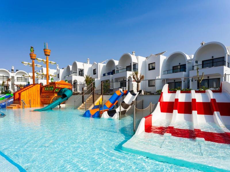 Großer bunter Wasserspielplatz mit Rutschen und angrenzenden weißen Bungalows unter blauem Himmel.