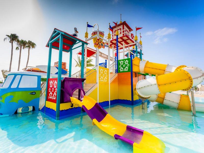 Buntes Wasserspiel mit Rutschen, Türmen und Palmen in klarem blauem Wasser unter blauem Himmel.