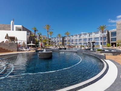 Modernes Hotel mit großem Pool, Sonnenliegen und Palmen unter klarem blauem Himmel.