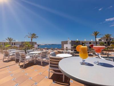 Terrasse d'hôtel ensoleillée avec tables, chaises et vue sur la piscine et la mer.