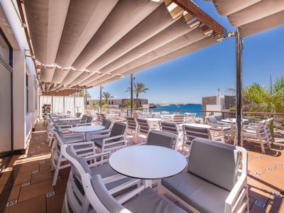 Terrasse mit Tischen und Stühlen, überdacht, mit Blick auf das Meer bei klarem Himmel.