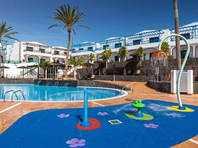 Bassin pour enfants avec jeux d'eau à côté d'une grande piscine et de plusieurs bâtiments aux accents bleus.
