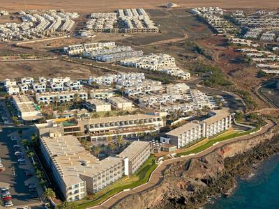 Vue d'un complexe hôtelier avec des bâtiments blancs sur une côte rocheuse et des montagnes en arrière-plan.